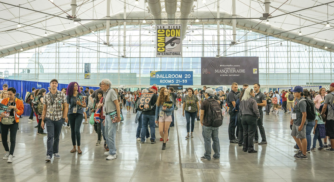 An image of a group of Comic-Con attendees walking through a large well-lit room with large windows in the background. A Comic-Con sign hangs in the center of the image.